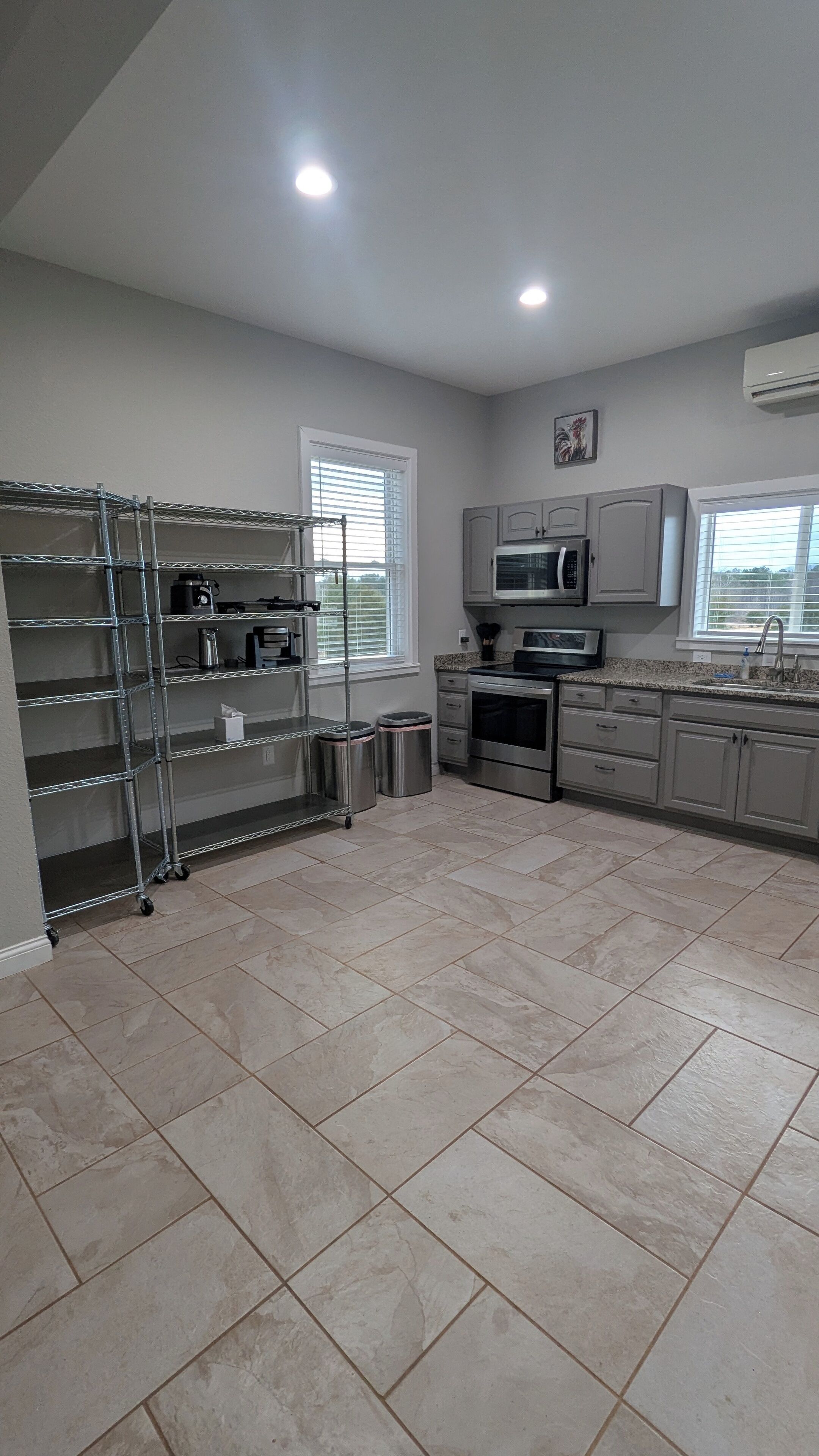 A kitchen with a stainless steel range hood, a microwave, and a dishwasher. There is a window with blinds, a stainless steel shelf unit, and a tiled floor.