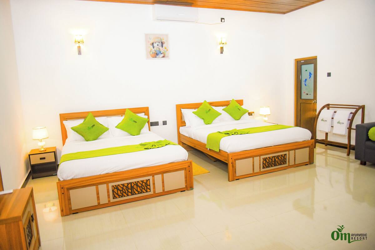 A hotel room with two wooden beds, each with green pillows and white bedding. There are bedside lamps, a small table, and a wall-mounted air conditioner.