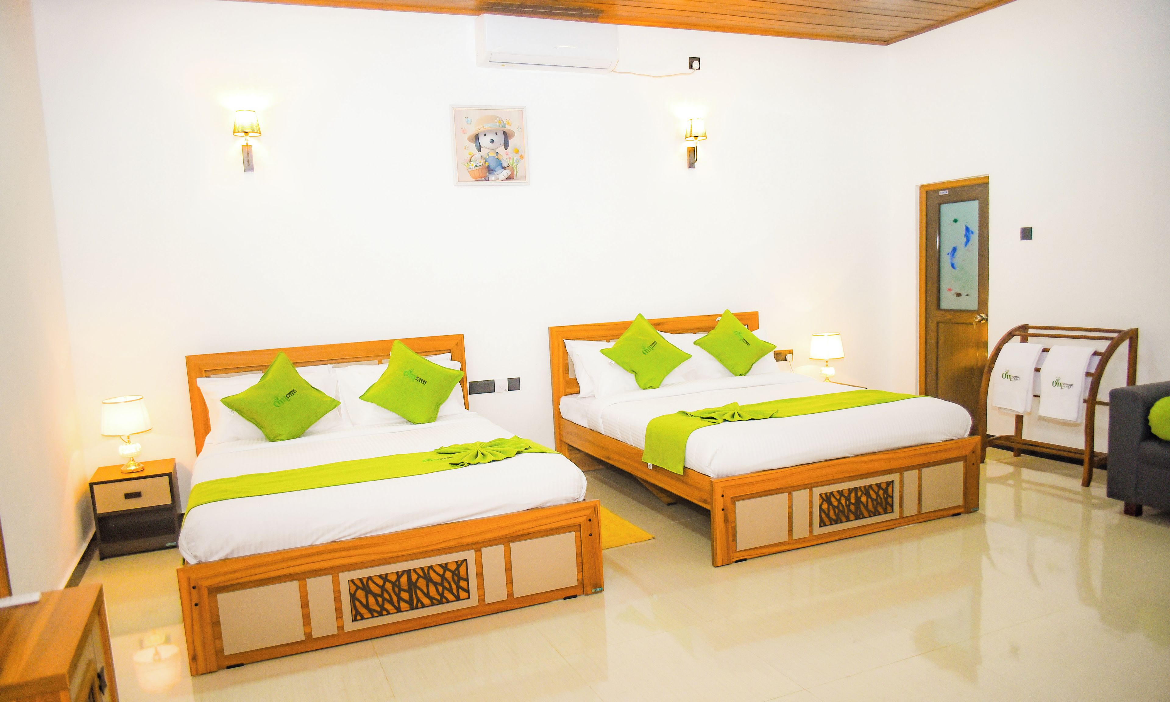 A hotel room with two wooden beds, each with green pillows and white bedding. There are bedside lamps, a small table, and a wall-mounted air conditioner.