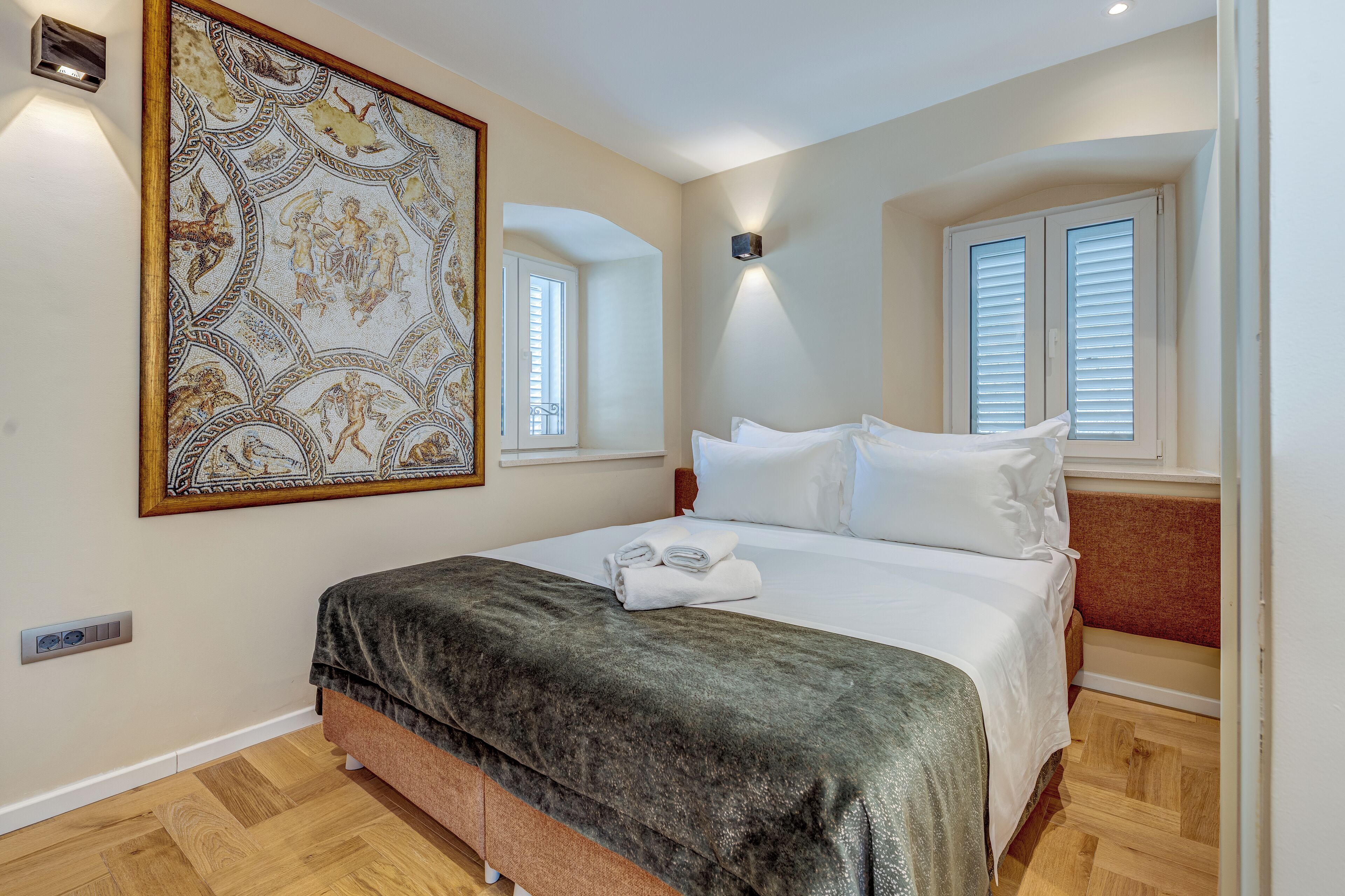 Une chambre avec un grand lit, un élément décoratif mural, une fenêtre avec des stores et un luminaire fixé au mur.