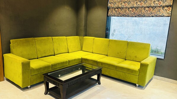 A bright yellow sectional sofa with a glass coffee table in front, positioned against a dark wall with a window and patterned curtain. - Hotel Grand Avenue (Lucknow)