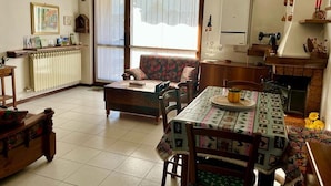 Een woonkamer met een bank, een eettafel met een tafelkleed met patroon, een radiator en een open haard.