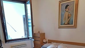 Een netjes opgemaakt bed met een blauw-beige sprei, een houten nachtkastje met een lamp, een schilderij aan de muur en een raam met gordijnen.
