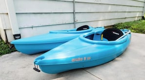 Two blue kayaks with the text "aruba 8 ss" on one of them, placed on a concrete surface in front of a white building.