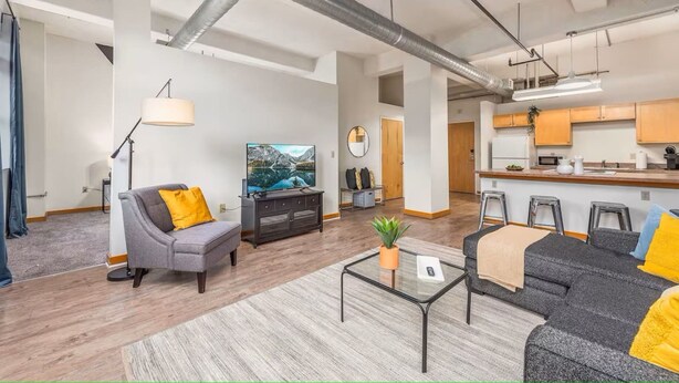 Ein modernes Wohnzimmer mit Sofa, Couchtisch und Fernseher. Es gibt eine Küchenzeile mit Barhocker und einem Esstisch.