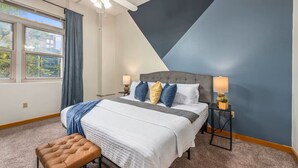 Ein Schlafzimmer mit einem Bett, zwei Nachttischen, einer Bank, einem Deckenventilator und einem Fenster mit Vorhängen.