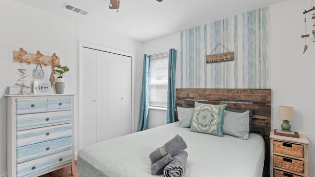 Une chambre à coucher avec un lit, une tête de lit en bois, une table de chevet, une commode, un ventilateur de plafond et un mur orné d’une inscription « RELAX ».