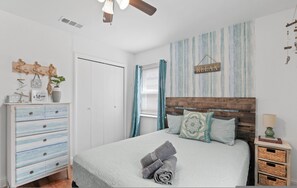 Ein Schlafzimmer mit Bett, Holz-Kopfteil, Nachttisch, Kommode, Deckenventilator und einer Wand mit dem Schriftzug 'RELAX'.