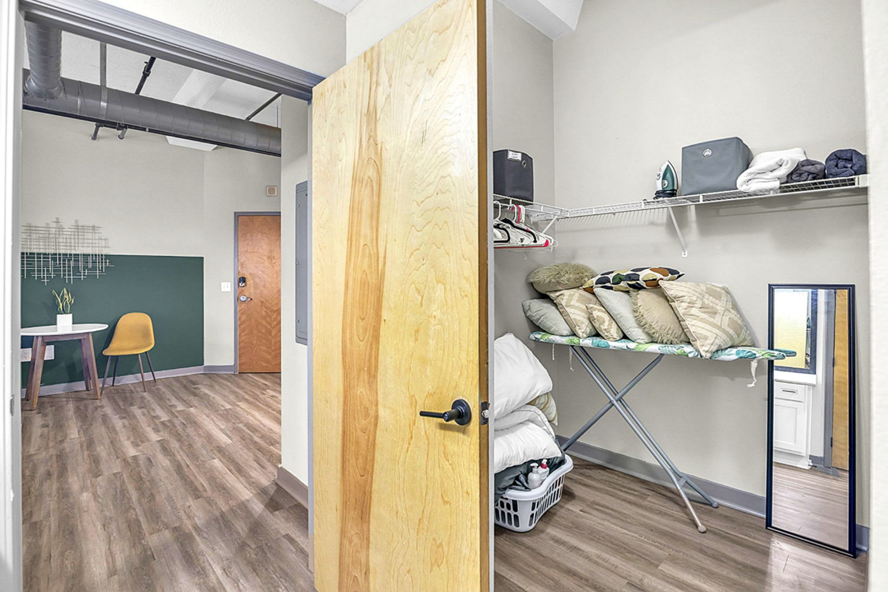 Una habitación con puerta de madera, una mesita con una silla, un estante con ropa y una cesta de lavandería.