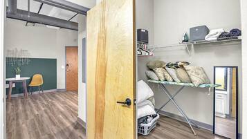 Una habitación con puerta de madera, una mesita con una silla, un estante con ropa y una cesta de lavandería.