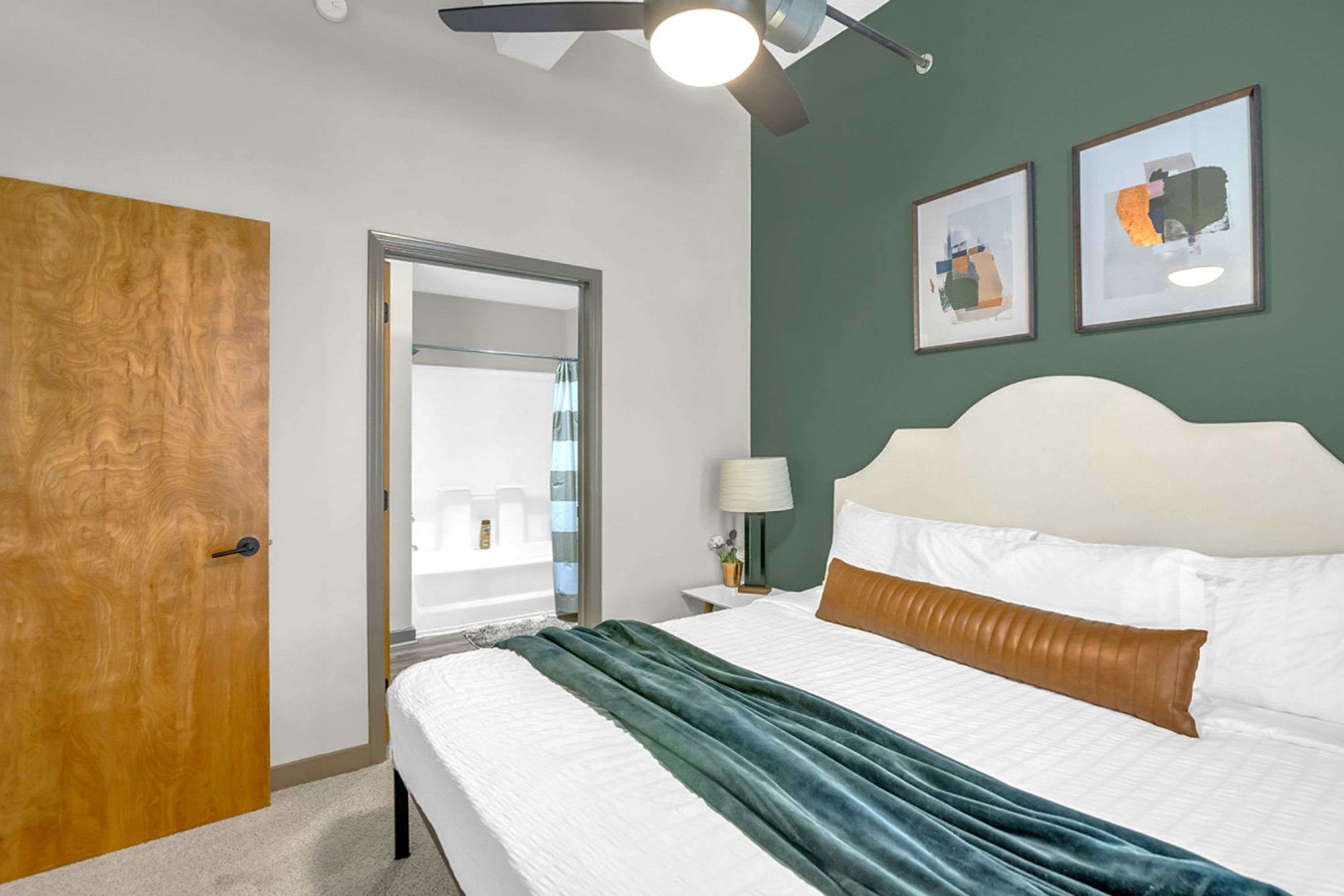 Una habitación de hotel con una cama, una puerta de madera, un ventilador de techo y dos cuadros en la pared.