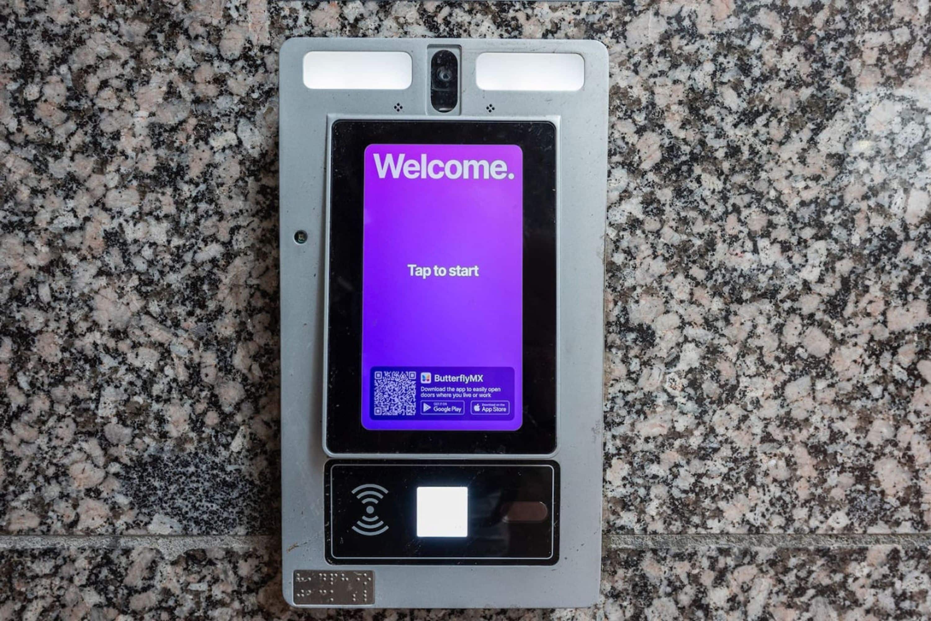 A digital kiosk with a purple screen displaying 'Welcome. Tap to start.' and a QR code for an app download.