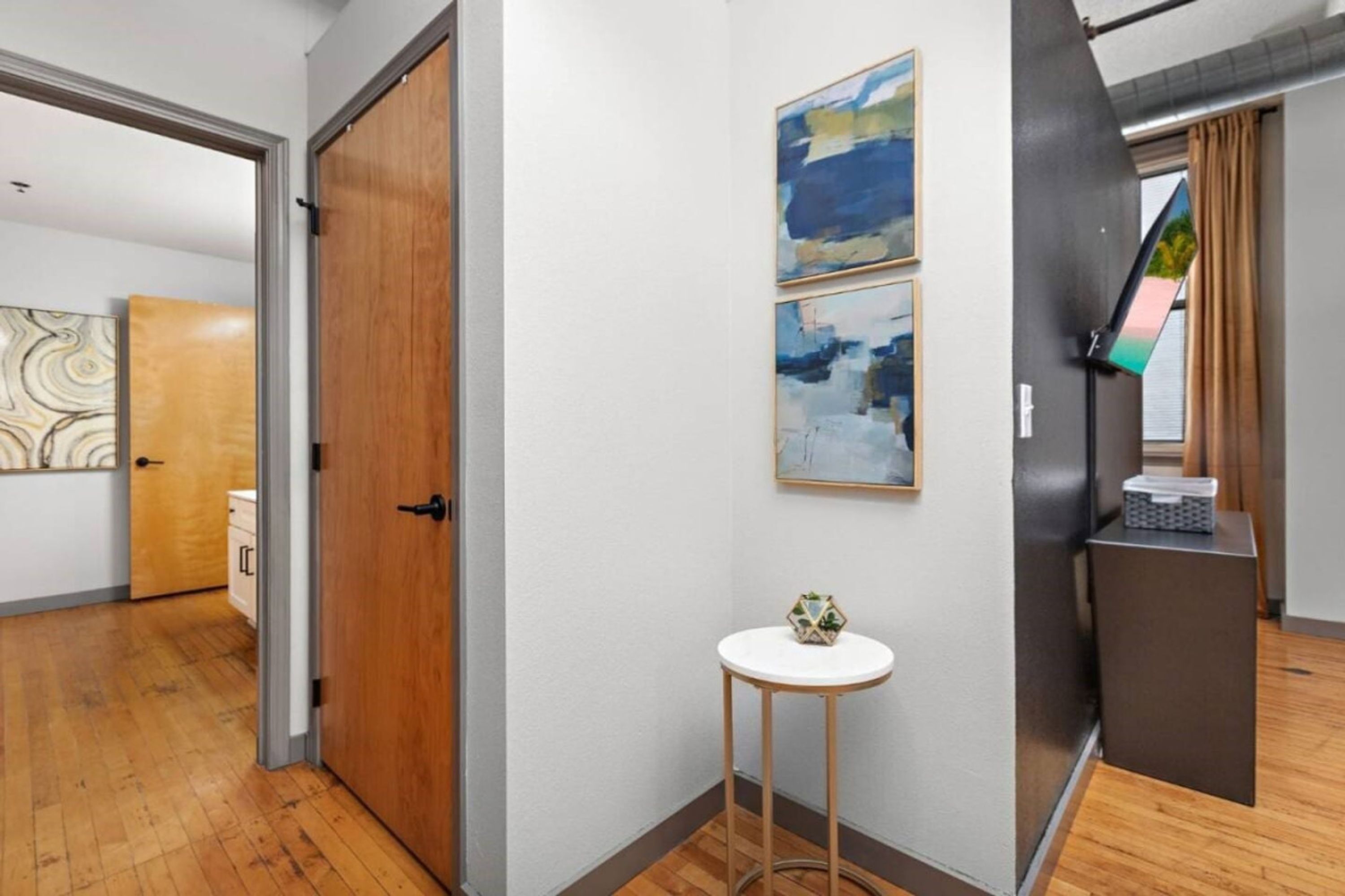 A hallway with a wooden floor, a white door, and a small white table with a decorative item. There are abstract paintings on the wall and a window with curtains.