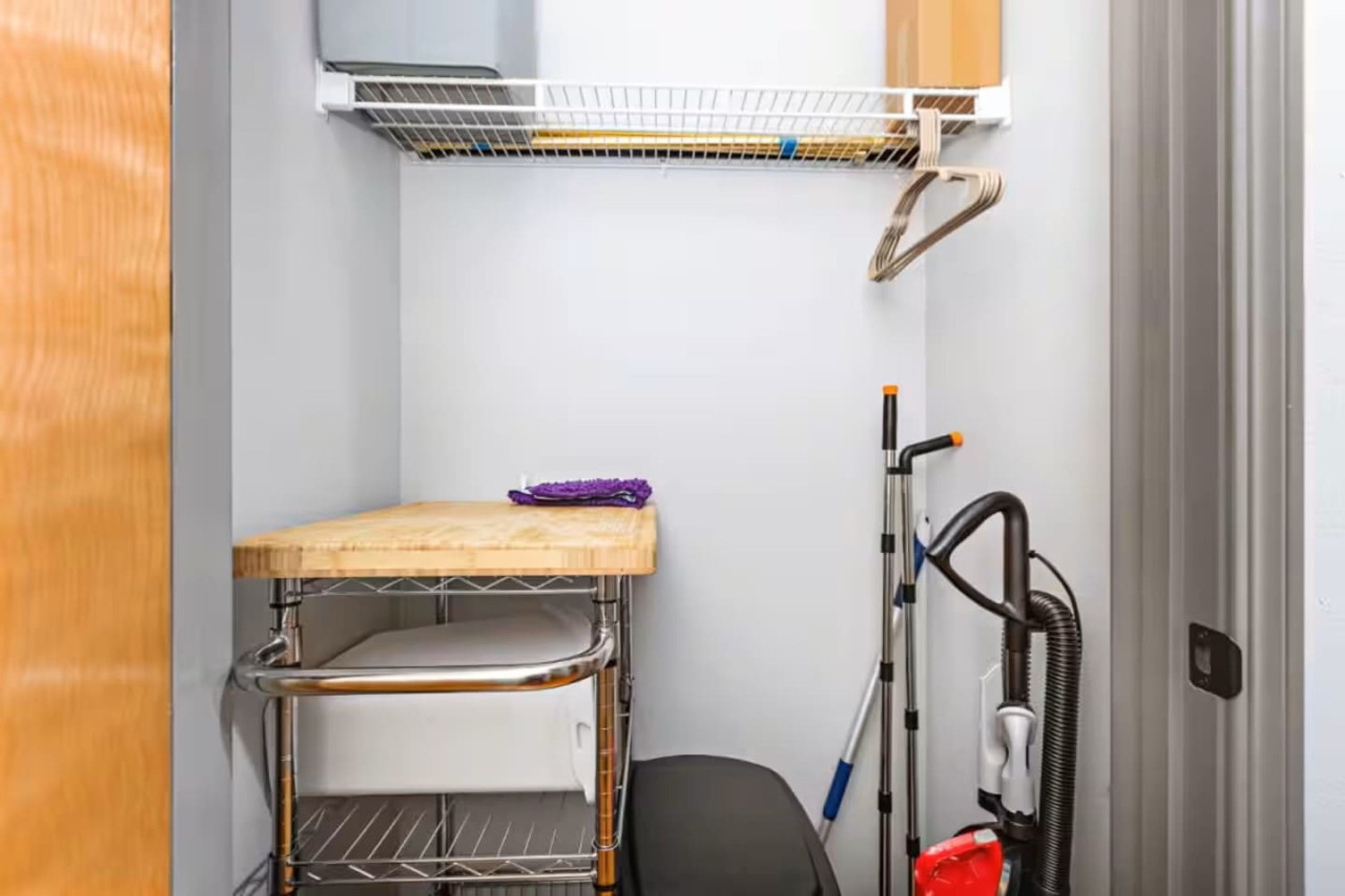 Quarto compacto com uma estante de metal, uma pequena mesa de madeira e um aspirador de pó.