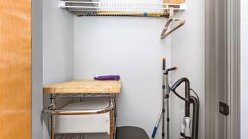 Quarto compacto com uma estante de metal, uma pequena mesa de madeira e um aspirador de pó.