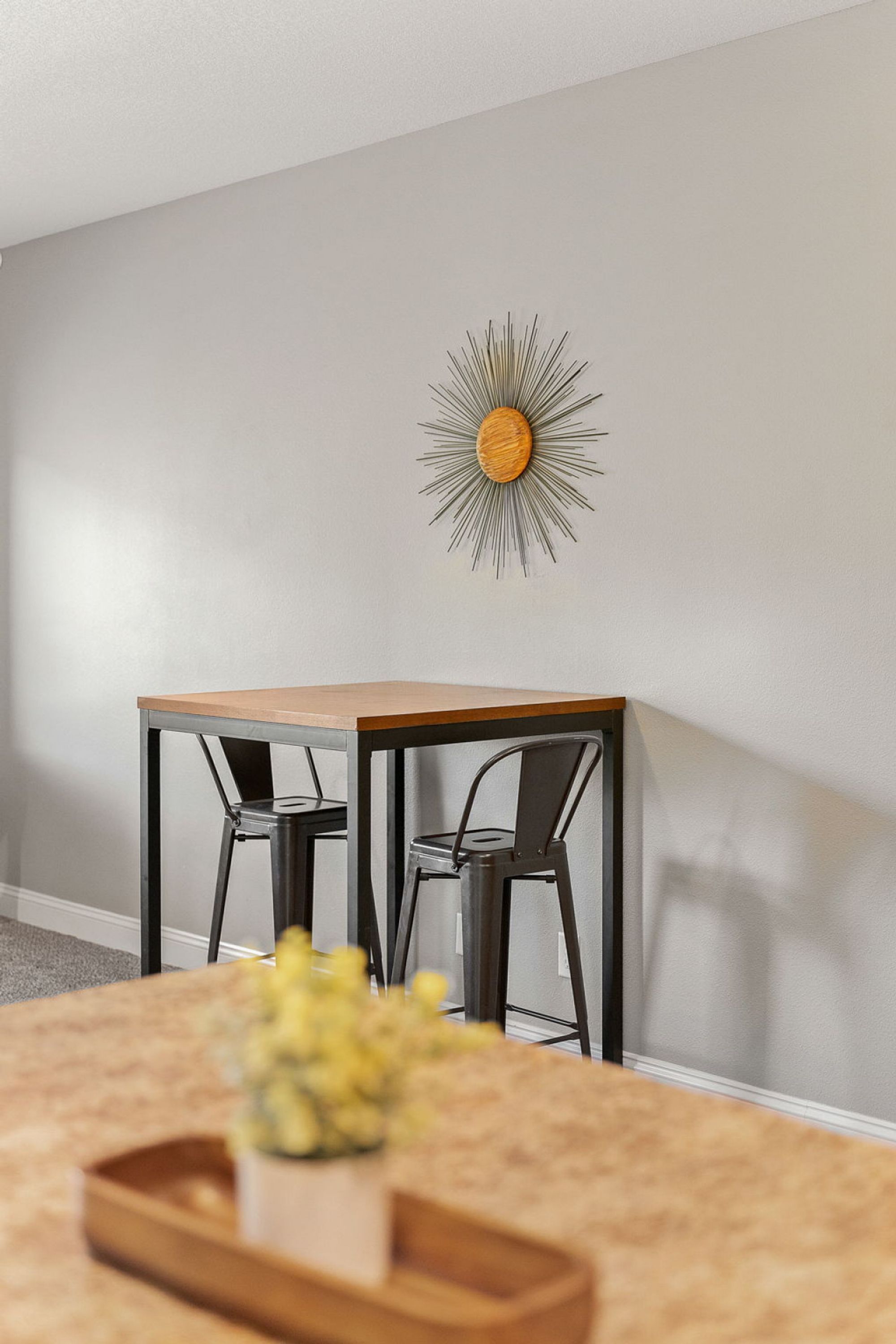 Zona pranzo con tavolo e due sedie, un’opera d’arte a forma di sole decorativo e una piccola pianta sul tavolo.