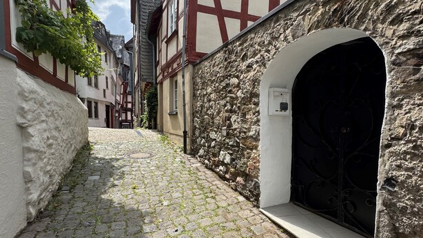 Calle adoquinada bordeada por edificios tradicionales con entramado de madera, una pared de piedra y una entrada en arco.