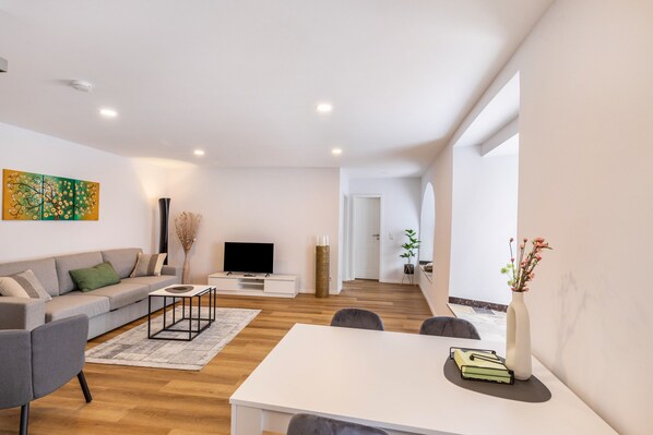 A modern living room with a sofa, coffee table, flat-screen TV, and a dining area with a table and chairs.
