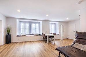 A spacious room with a dining table, chairs, a sofa, and large windows with curtains.