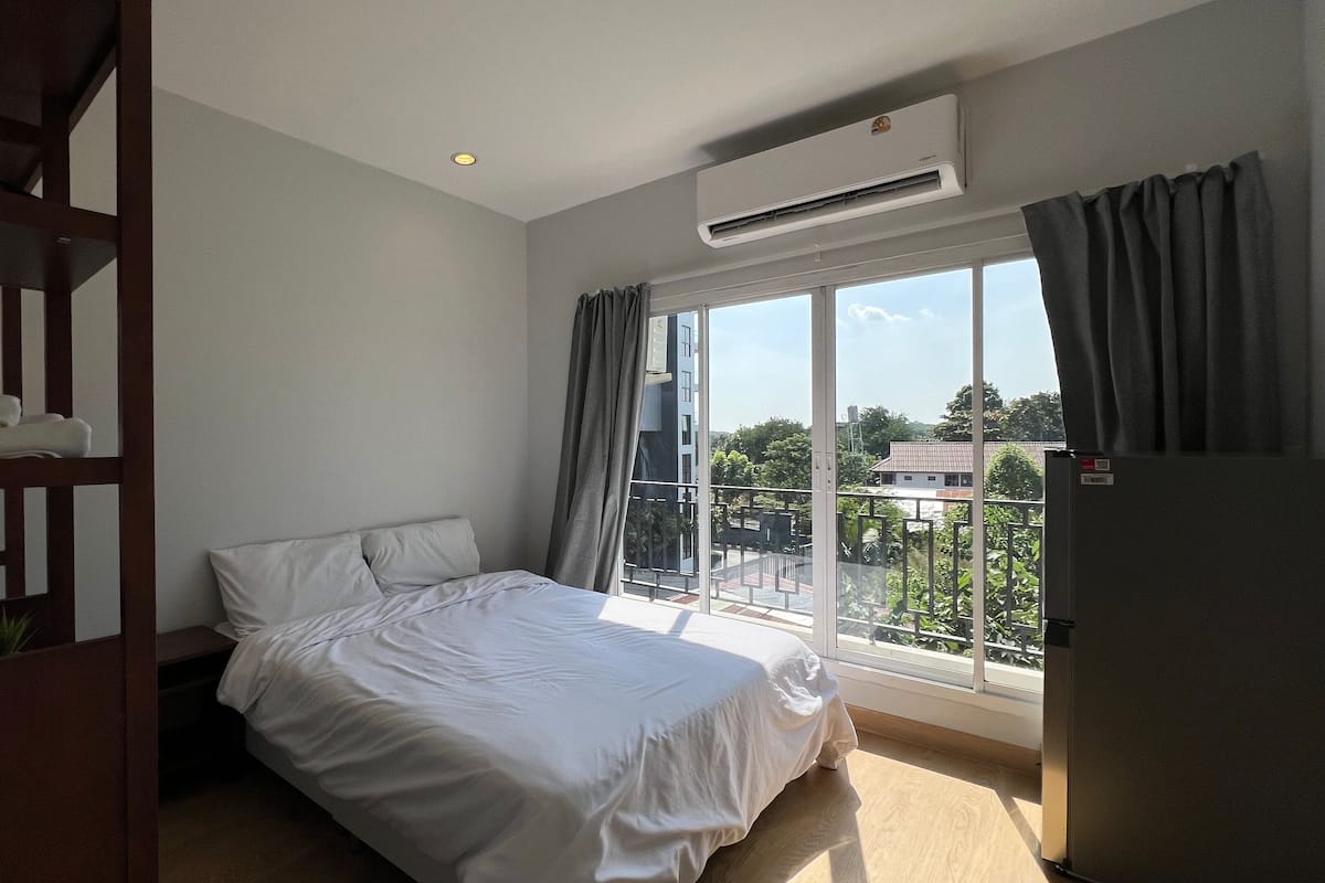 A hotel room with a bed, a desk with a lamp, an air conditioner, and a view of the outside.