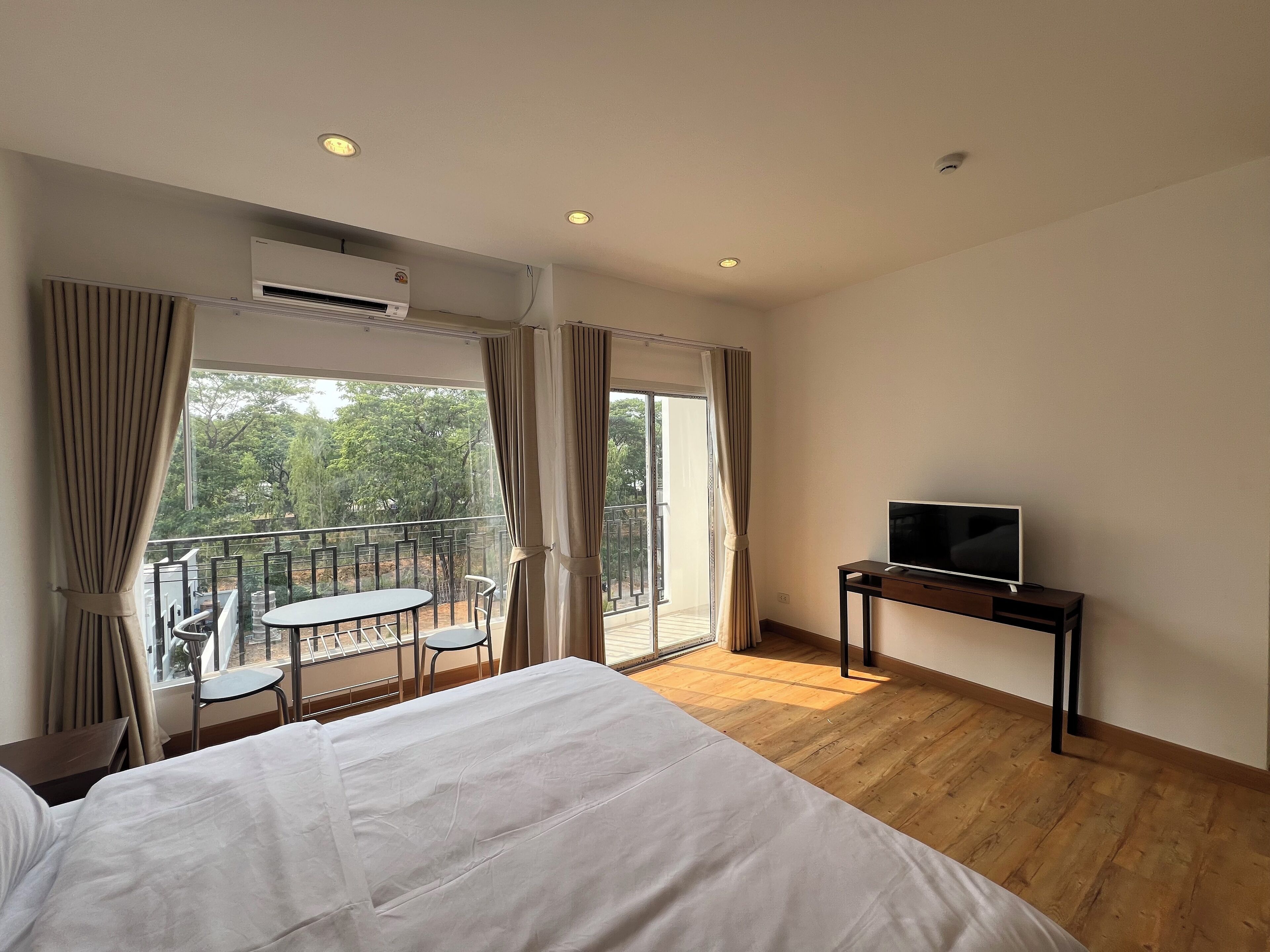 A hotel room with a bed, a TV on a stand, a balcony with a table and chairs, and an air conditioning unit.