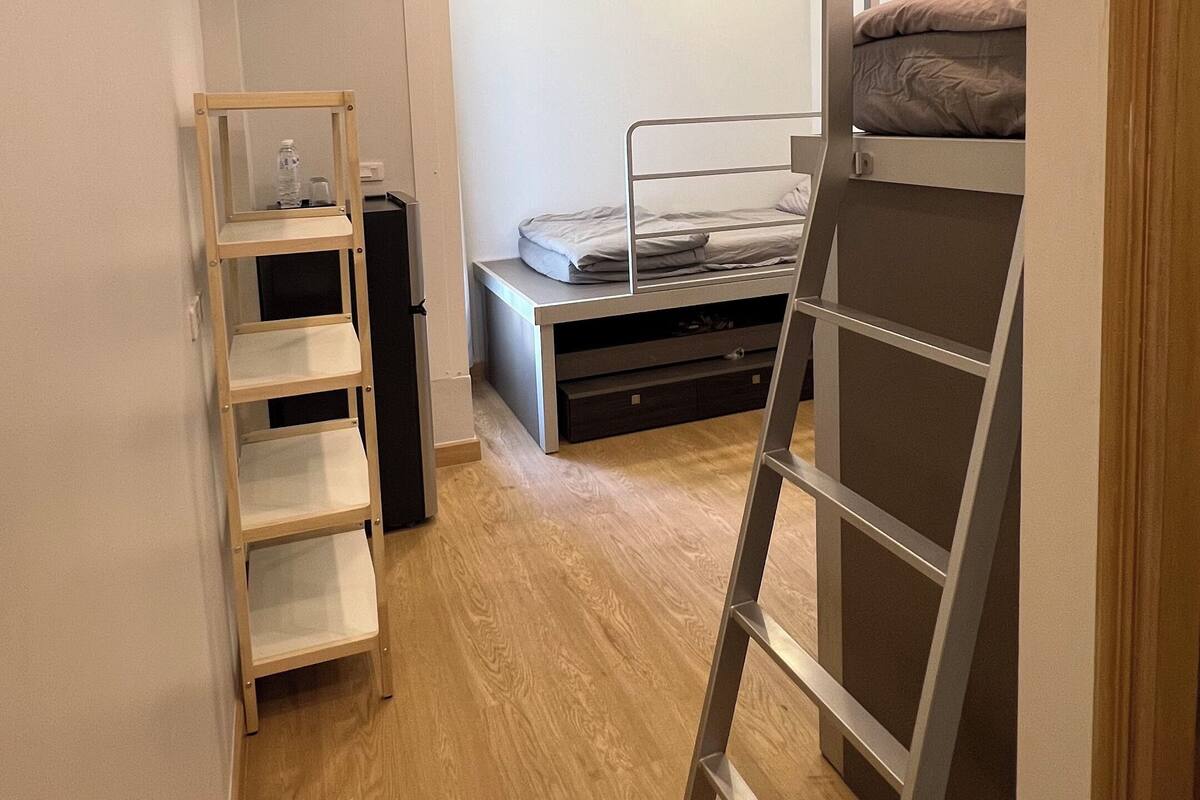 A compact room with a bunk bed, a ladder, a small shelf unit, and a ceiling-mounted air conditioning unit.