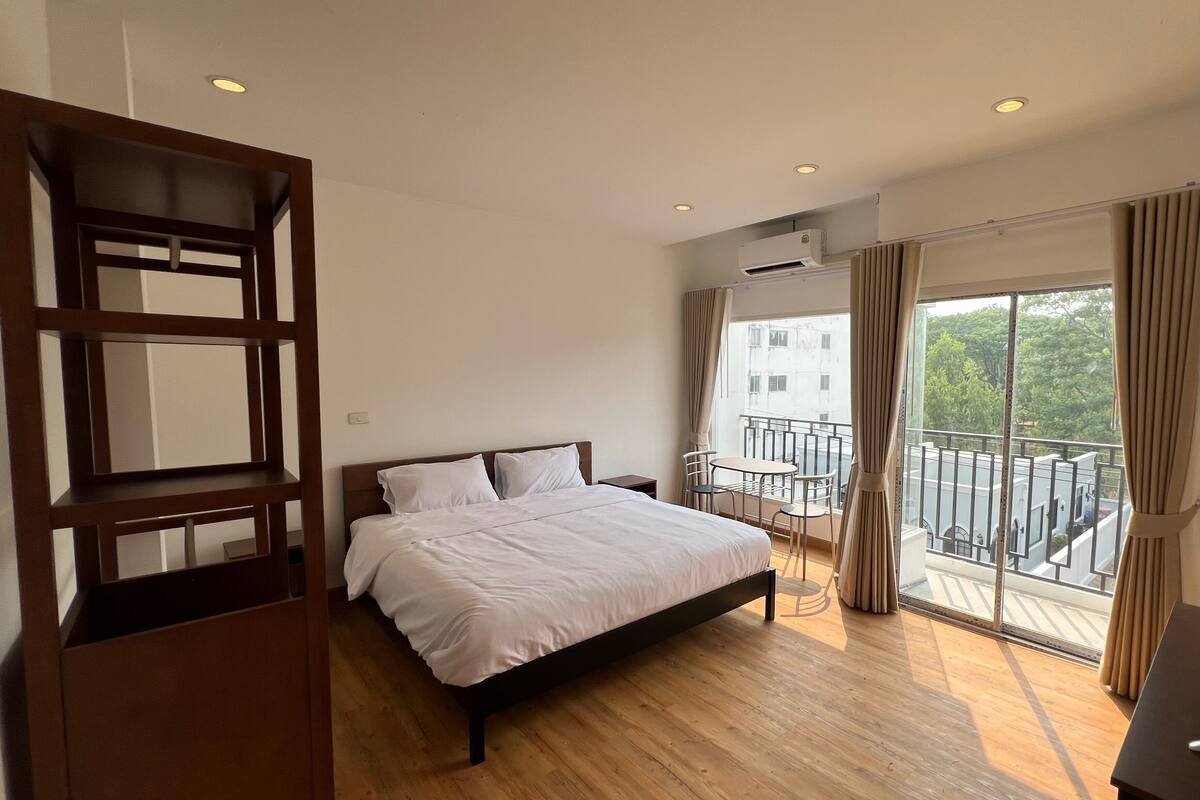 A bedroom with a bed, a wooden shelf, a balcony with a table and chairs, and an air conditioning unit.