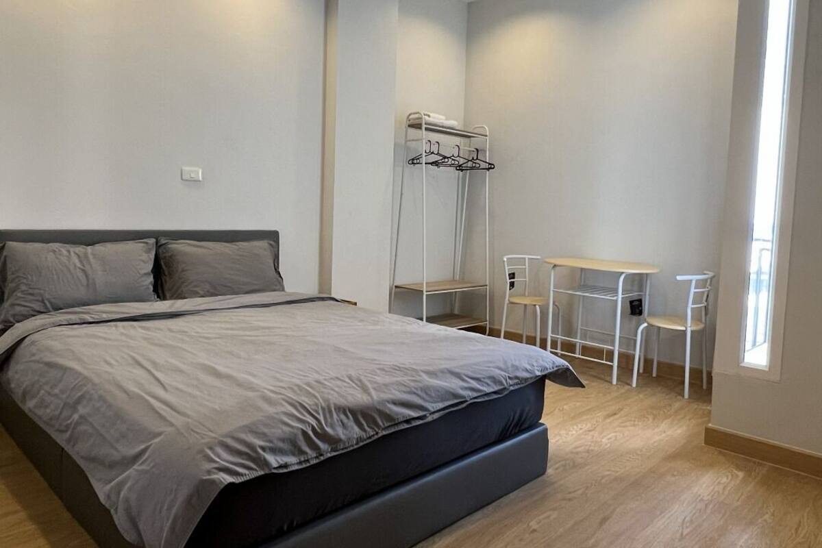 A modern bedroom with a large bed, a small table, and a chair, a coat rack, and a window providing natural light.