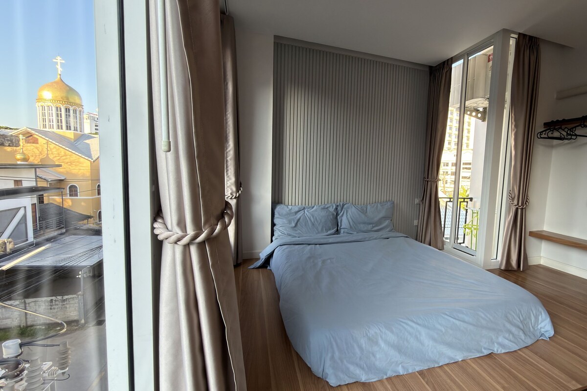 A bedroom with a bed, curtains, a window with a view of a building, and a wooden floor.