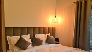 Un grand lit avec une tête de lit en bois, un luminaire suspendu et une vue sur l’extérieur par une fenêtre ornée de rideaux.