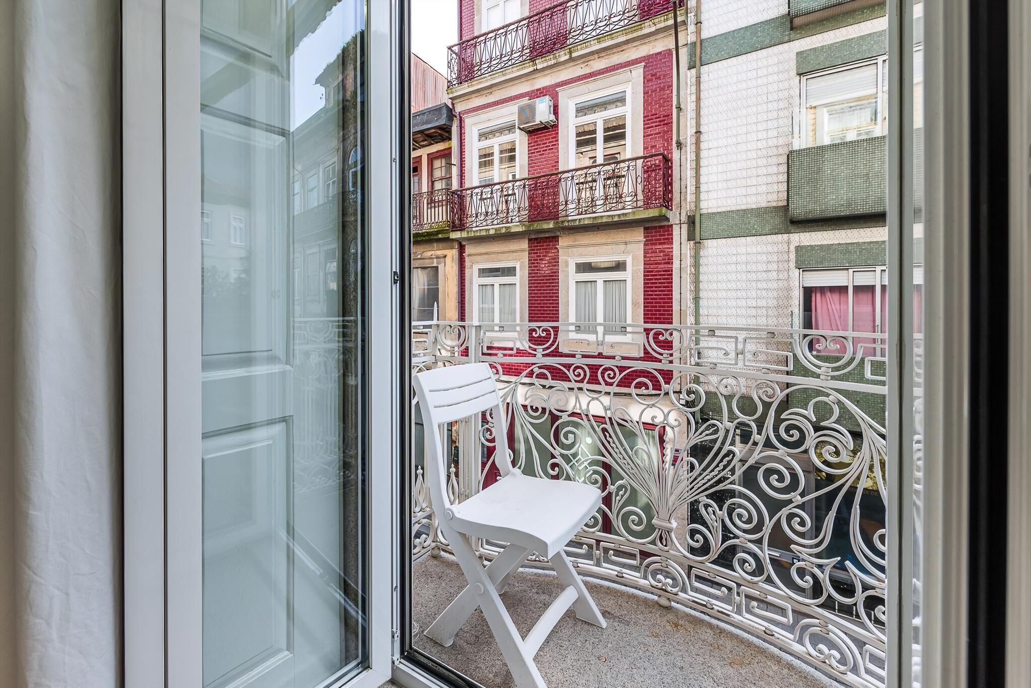 Un balcón con una silla blanca, una barandilla metálica y vistas a un edificio rojo y blanco.