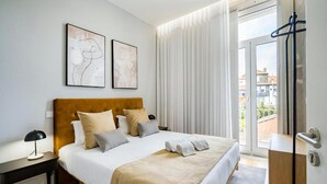 A hotel room with a bed, a nightstand, a desk, a chair, a window with curtains, and a view of buildings outside.