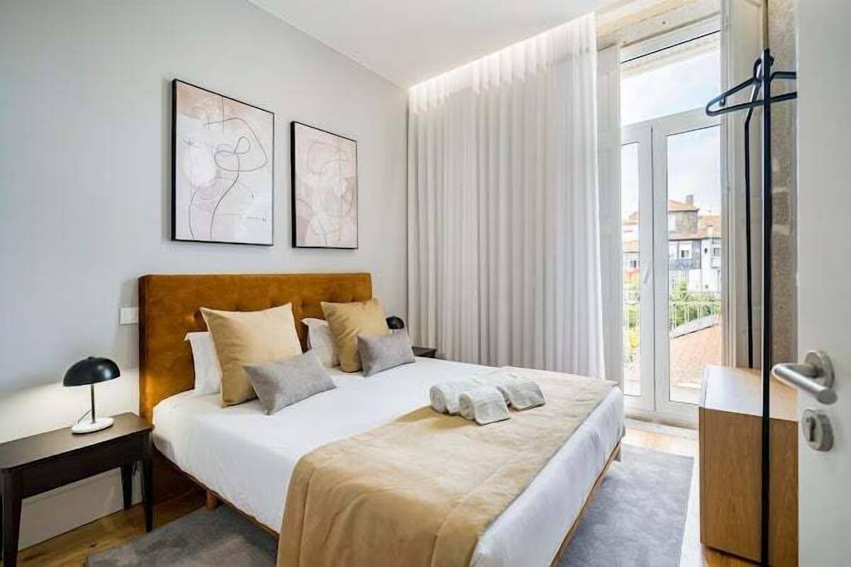 A modern hotel room with a large bed, a wooden headboard, two abstract wall art pieces, a bedside table with a lamp, and a view through a glass door to the outside.