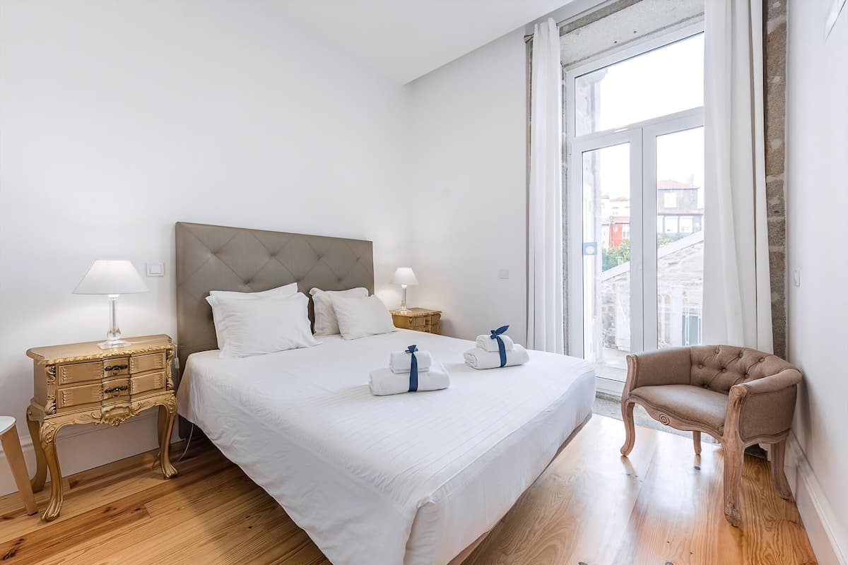 A bedroom with a large bed, two bedside tables, a chair, and a window with curtains.