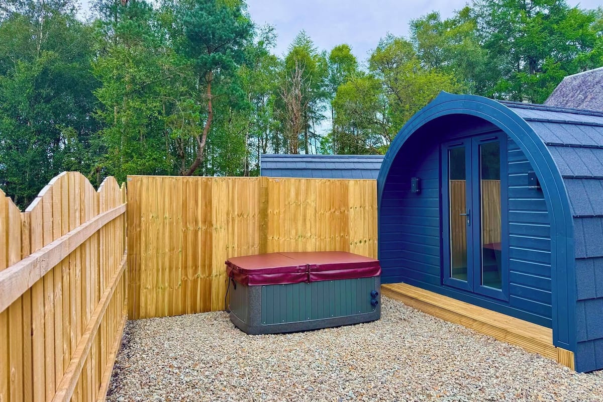 Un petit espace extérieur clos, délimité par une clôture en bois, comprenant un jacuzzi et un bâtiment bleu doté d’un toit incurvé.
