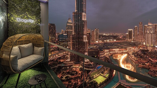 A high-rise room with a view of a city skyline, a green wall with a logo, a wicker sofa, and a small table. - Gogoto1 2BR Burj Khalifa, Fountain View Dubai Mall (Downtown Dubai)
