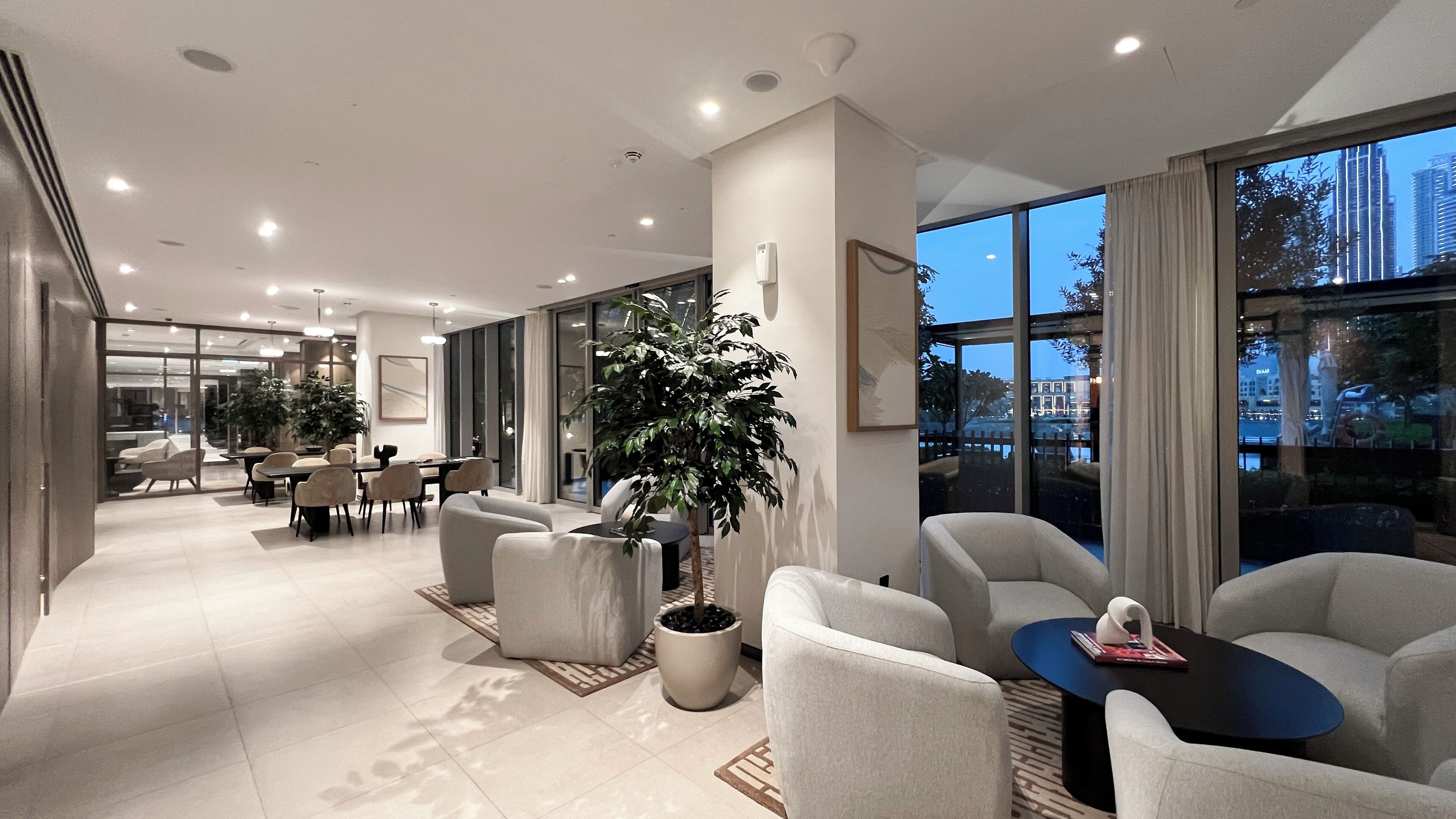 Un hall d’hôtel moderne doté d’un espace repas, de différents coins salon et de grandes fenêtres offrant une vue sur la ville.