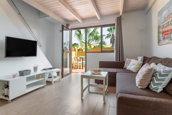 A modern living room with a flat-screen TV, a white console table, a brown sofa with patterned cushions, and a view of palm trees through large windows.