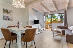 A dining area with a white table and brown chairs, a TV mounted on the wall, and a view of palm trees through the open door. - Marvin Home in Corralejo: Renovated Apartment with Garden View (Corralejo)