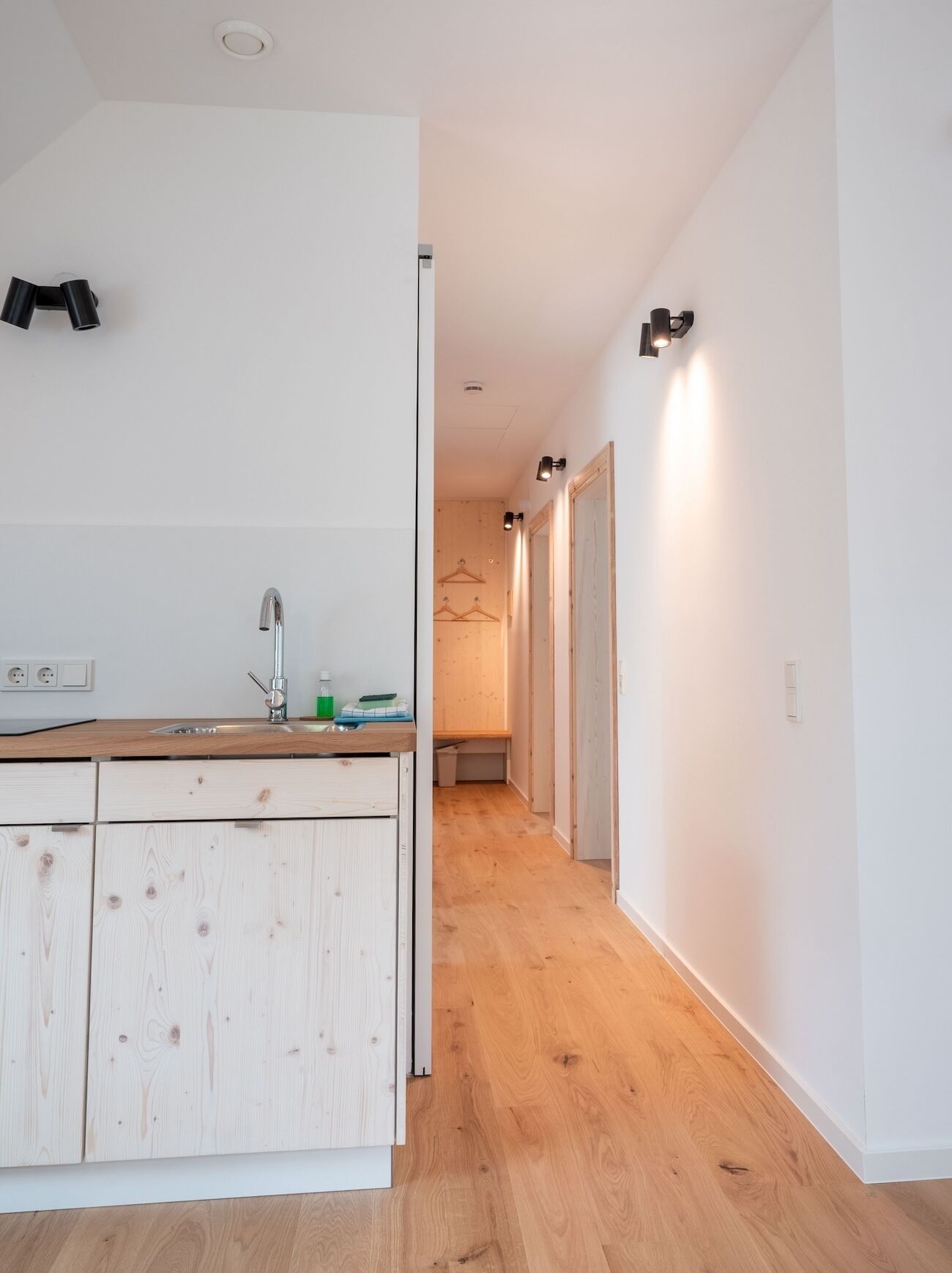 Uma cozinha moderna com bancada de madeira, uma pia e um armário. Há um corredor com piso de madeira e uma porta que dá para outro cômodo.