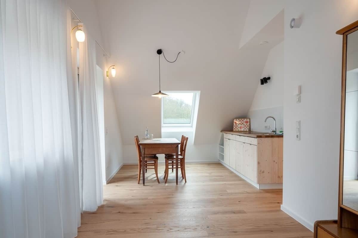 Une pièce comprenant une table à manger, des chaises, un luminaire suspendu, une fenêtre et un coin cuisine.