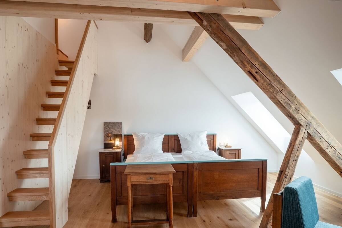 Une chambre à coucher aménagée dans un espace en hauteur, avec un escalier en bois, un lit avec une literie blanche, une petite table et une chaise.