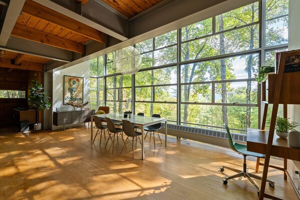 A spacious room with large windows, a dining table, chairs, a sofa, and a bookshelf.