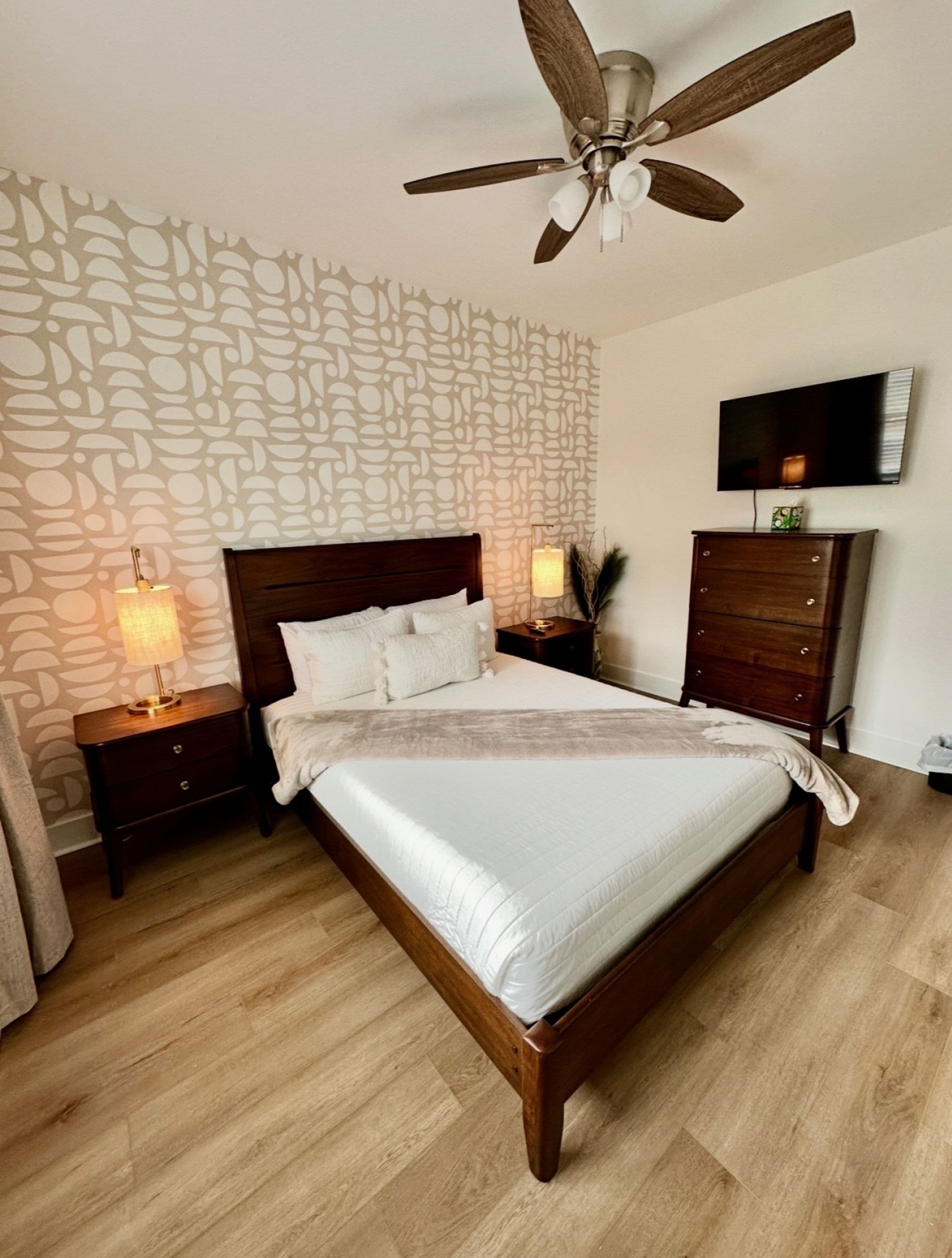 A bedroom with a wooden bed, a ceiling fan, a TV on the wall, and a patterned wall.