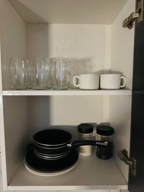 A kitchen cabinet with glassware, white mugs, and black cookware.