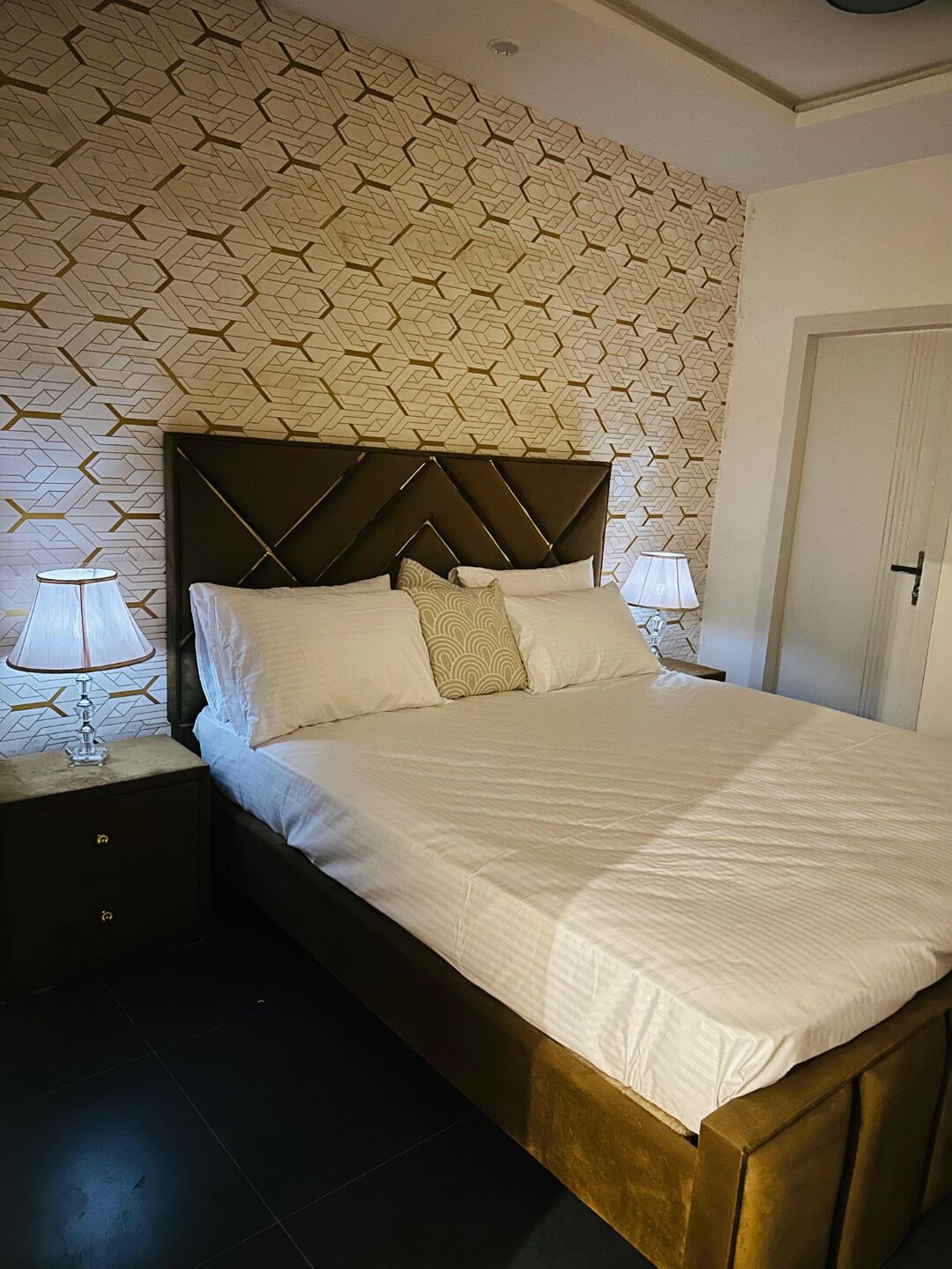 En snyggt bäddad säng med ett mönstrat sänggavel, två sänglampor och en dörr till höger.