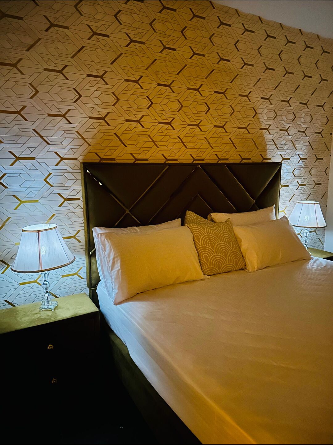 A neatly made bed with a geometric patterned headboard, a bedside lamp, and a wall with a similar geometric pattern.