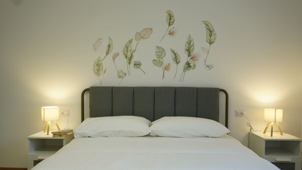 A neatly made bed with white linens, flanked by two bedside tables with lamps, and a wall-mounted artwork of leaves. - Grimani Suite (Milan)