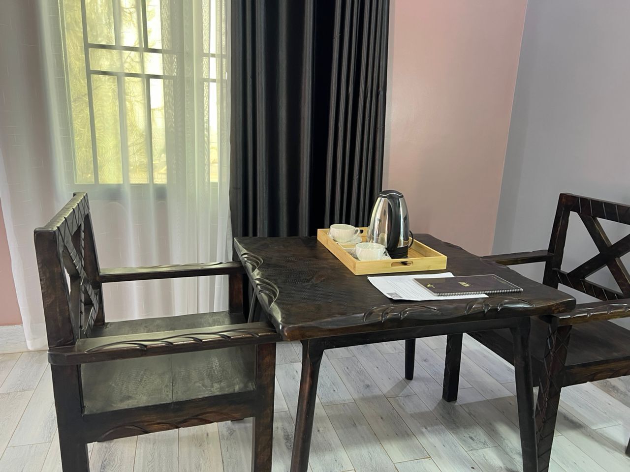 Uma área de refeições com uma mesa de madeira escura, duas cadeiras, um tabuleiro com chávenas e uma chaleira, e uma janela com cortinas.