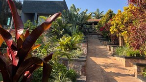 A pathway leading to a building with a dark roof, surrounded by various tropical plants and a stone wall. - REBERO RESORT  (Kigali)